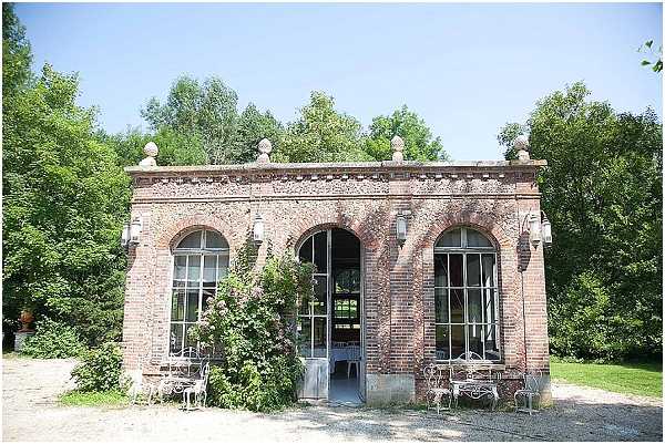 Exterior shot of a 19th-century French brick orangery or pavilion building featuring three large arched windows with tall glass doors, decorative stone ball finials along the roofline, and ornate brick detailing. The entrance is flanked by climbing plants and white wrought-iron garden chairs and tables are arranged outside on a gravel courtyard. The building appears to be set up as a wedding reception or cocktail hour space, with a glimpse of interior décor visible through the open central door. Wide establishing shot taken in bright midday light. Potential venue feature image.