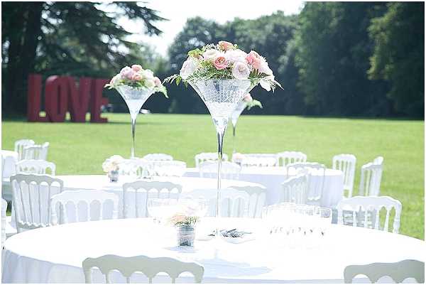 An outdoor wedding reception setup on a large green lawn, showing round tables covered in white linen cloths surrounded by white Napoléon chairs. The foreground table features a tall martini-glass vase centerpiece holding a cluster of blush pink and ivory roses with greenery, with a small matching arrangement in a silver pot alongside place card holders. Additional identically styled tables are visible in the background. A large red freestanding 'LOVE' letter installation is placed on the lawn in the background among mature trees. The overall decor palette is white and blush pink with silver accents, conveying a classic romantic theme. Wide shot taken from a slightly elevated angle.