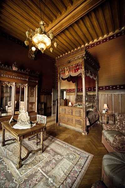 This image shows an ornate interior bedroom suite, likely within a historic French chateau, featuring a large carved wooden four-poster canopy bed with deep red and burgundy drapery and fringe detailing. The room includes a beamed wooden ceiling, an antique chandelier with amber glass shades, heavily carved wooden wall paneling and armoire, a writing desk with an upholstered chair, and a large decorative area rug in muted floral tones. No people are present in the frame; this appears to be a wide interior shot showcasing the room's architectural and decorative features. Potential venue feature image.