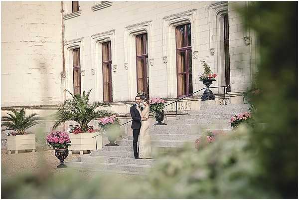 A couple poses together on the stone entrance steps of a French chateau during what appears to be a wedding portrait session. The bride wears a fitted ivory gown and the groom is dressed in a dark navy suit. The chateau facade features tall arched windows with dark wood frames, and the entrance is decorated with large ornate black iron urns and white rectangular planters filled with pink and red flowering plants. The shot is a wide environmental portrait framed through foreground foliage, positioning the couple centrally against the grand building exterior. Potential venue feature image.