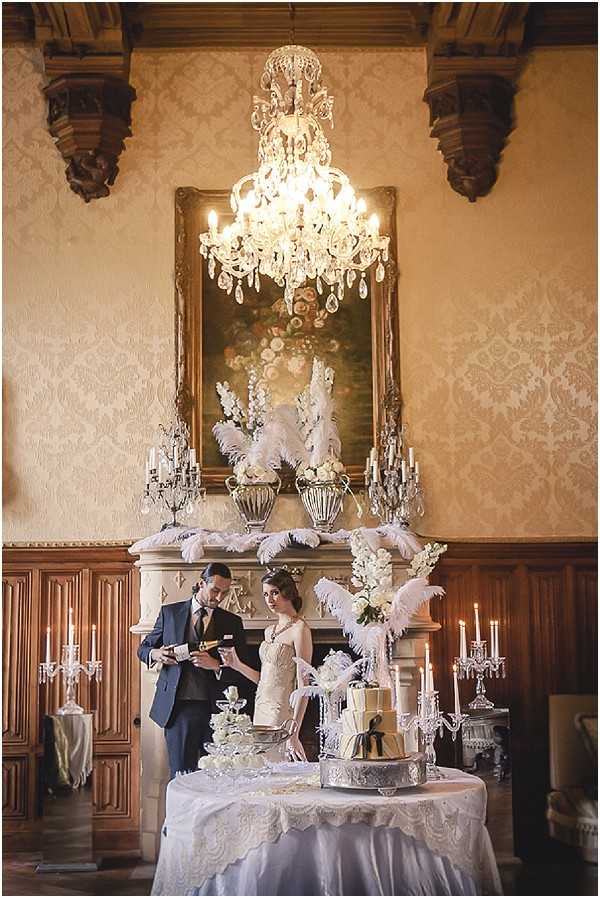 A couple poses at a cake cutting moment inside a grand chateau ballroom with heavily ornate damask-patterned walls, carved dark wood paneling, and a large crystal chandelier overhead. The groom wears a navy suit and is opening a bottle of champagne, while the bride wears a fitted ivory lace gown with a pearl necklace. The wedding cake table is draped in a white lace-trimmed cloth and holds a tiered gold and white cake surrounded by crystal candelabras with lit white candles. The mantelpiece behind them is styled with white ostrich feather arrangements, white floral urns, and silver candelabras, beneath a gilt-framed oil painting. The overall decor theme is 1920s Great Gatsby-inspired, with a palette of ivory, white, gold, and silver. Wide portrait shot. Potential venue feature image.