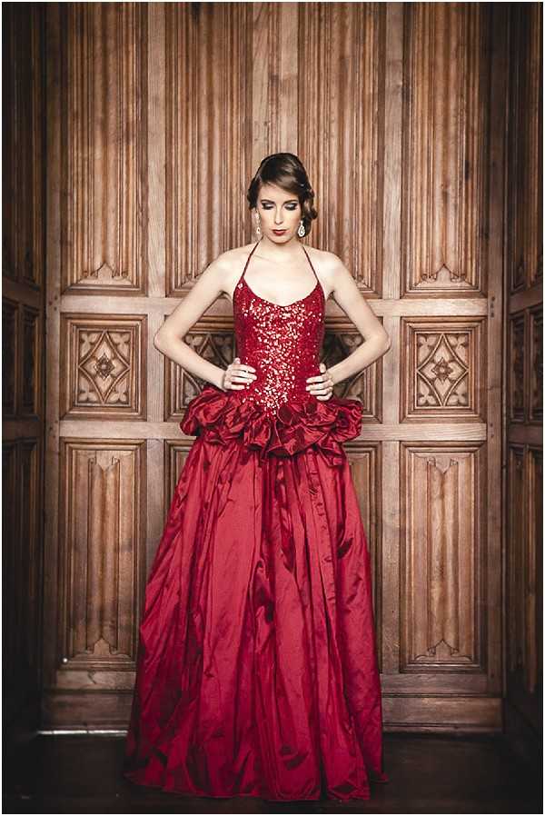 A portrait-style editorial or styled shoot image featuring a woman in a deep crimson red halter-neck ball gown with a heavily sequined and beaded bodice and a voluminous ruched taffeta skirt with a dramatic peplum detail at the waist. She stands with hands on hips, looking downward, posed against ornately carved dark wood paneled doors with geometric and floral relief detailing. Her hair is styled in an updo and she wears drop earrings. The overall styling is dramatic and formal, with a bold color-forward aesthetic. Full-length portrait composition shot indoors.