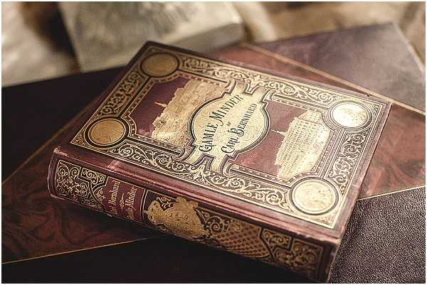 Close-up detail shot of an antique hardcover book with a deep burgundy and gold decorative cover, featuring ornate gilded scrollwork, medallion corners, and an embossed architectural vignette at the center. The book appears to be a vintage Scandinavian title reading 'Gamle Minder' by 'Chr. Bernhard,' used as a decorative prop or personal heirloom incorporated into the wedding styling. It rests on a marbled surface alongside what appears to be a dark leather-bound item, with soft fabric visible in the background. The styling suggests a classic or vintage-inspired wedding aesthetic, with the prop adding a personal or literary detail to the decor.