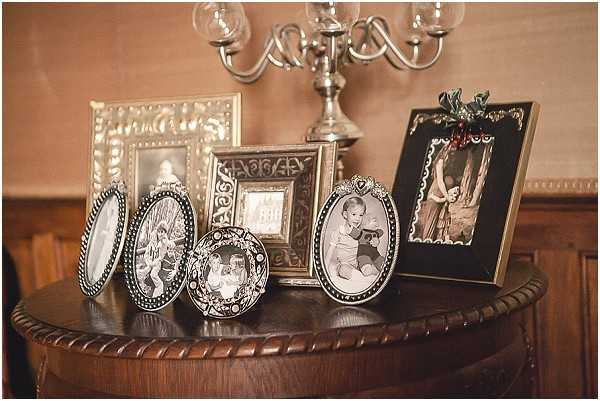 A close-up detail shot of a collection of ornate photo frames arranged on a dark wooden side table, likely used as a wedding decor display featuring family photographs. The frames vary in shape — oval, round, rectangular, and square — with silver, gold, and black finishes featuring decorative embossed detailing. The photographs inside appear to be vintage or childhood family images in black and white and sepia tones. A silver candelabra is visible in the background, set against warm-toned wood-paneled walls, suggesting an interior venue with a classic, traditional decor style.