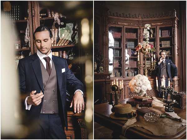 A diptych of groom portraits taken indoors in a richly decorated library or study, featuring dark carved wooden bookshelves lined with leather-bound books. On the left, a close-up portrait shows the groom adjusting his suit jacket; he wears a navy blue suit with a taupe double-breasted waistcoat, a brown tie, and a white pocket square. On the right, a wider shot shows the groom standing in the background while the foreground features a desk styled with a large arrangement of cream garden roses and white blooms, brass candelabras with lit candles, a vintage telephone, a straw boater hat, and aged books, creating a classic vintage editorial aesthetic. The overall styling theme is vintage or Great Gatsby-inspired, with warm amber tones and moody lighting throughout both frames.