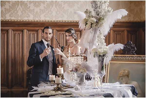 A styled shoot portrait of a couple posed at a decorated table inside a chateau or manor house, featuring dark wood paneling, ornate gold-framed mirrors, and textured wallpaper. The groom wears a navy suit with a grey waistcoat and holds a cigarette holder, while the bride wears a beaded, low-cut 1920s Great Gatsby-style gown with pearl jewelry and finger-waved hair. The table decor features large white ostrich feather arrangements with white orchids and roses, brass candlesticks with lit ivory candles, crystal glassware, and a black-and-white patterned table runner, all styled in a 1920s Art Deco theme with a predominantly white, gold, and black palette. The shot is a medium-wide portrait framing both subjects from the waist up against the richly decorated interior backdrop.