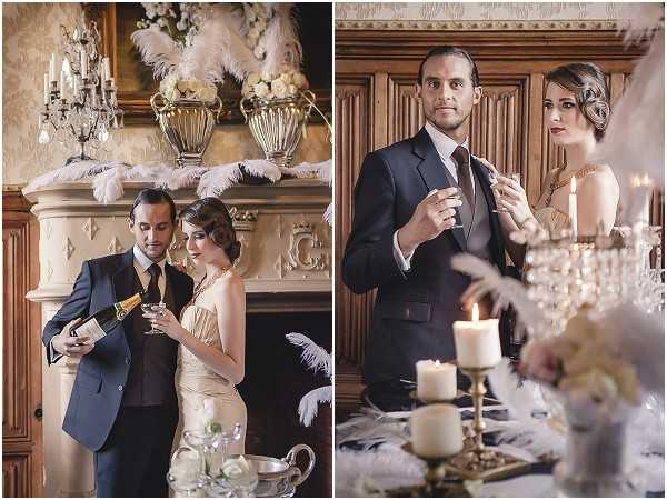 A styled wedding shoot featuring a couple posed indoors in two side-by-side portrait shots, both set within what appears to be a chateau or historic manor interior with ornate wood paneling, a carved stone fireplace, and a crystal chandelier. The groom wears a dark navy suit with a tie, while the bride wears a gold/champagne slip-style dress with a pearl or gold necklace and a finger-wave 1920s Art Deco hairstyle with dark berry lip color. In the left image, the groom pours champagne into the bride's glass in front of the fireplace, which is decorated with large white feather plumes and ivory floral arrangements in silver urns. In the right image, the couple holds champagne glasses near a table styled with gold candelabras, white taper candles, scattered white feathers, and crystal glassware, reinforcing the 1920s Great Gatsby-inspired theme. The overall decor palette is ivory, gold, and champagne with feather and crystal accents throughout.