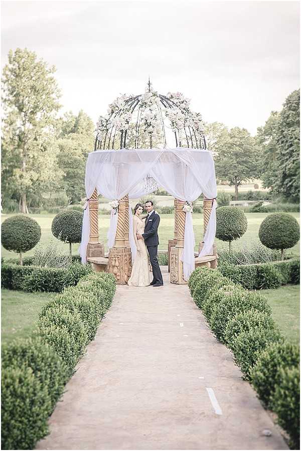 A couple poses together for a portrait in a formal outdoor garden, standing beneath an ornate wrought-iron domed gazebo structure decorated with white and blush floral arrangements at the top and white draped fabric panels on the sides. The bride wears a fitted, long-sleeved lace gown in a champagne-ivory tone, while the groom is dressed in a dark navy suit. The setting features a manicured formal garden with neatly clipped spherical topiary bushes, low boxwood hedging, and a straight gravel pathway leading up to the gazebo. The composition is a full-length wide portrait shot taken from the end of the pathway, centering the couple symmetrically within the structure and garden layout. Potential venue feature image.