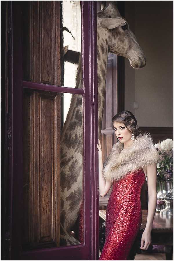 A bridal editorial or styled shoot portrait featuring a woman in a floor-length red sequined gown with a fur stole wrap, posed beside a dark burgundy-painted doorframe inside what appears to be an ornate interior room. She has a 1920s-inspired finger-wave hairstyle, bold red lips, and drop crystal earrings, suggesting a Art Deco styling theme. A taxidermied giraffe is visible through the glass panes of the door behind her, indicating an eclectic, maximalist interior décor setting, likely a chateau or manor house with hunting-lodge style decoration. In the background to the right, a floral arrangement in lavender and white tones and glassware are visible on a dark wood surface. The shot is a medium portrait with moody, warm ambient lighting.