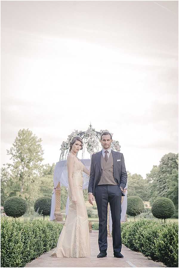 A couple poses together in a formal outdoor portrait in a manicured French formal garden, standing on a paved pathway flanked by clipped box hedging and topiary spheres. The bride wears a champagne-gold fitted lace gown with a low neckline and slight train, while the groom wears a charcoal suit with a brown double-breasted waistcoat and a dusty mauve tie. Behind them stands a wrought-iron gazebo or ceremony arch draped with sheer white fabric and decorated with white and soft green floral arrangements. The image has a soft, faded, high-key editing style with muted tones and low contrast, giving it a pale, washed-out finish. Full-length portrait shot.