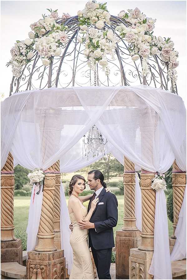 A couple poses together in a portrait shot beneath an ornate wrought-iron gazebo structure in an outdoor garden setting. The gazebo is draped with white sheer fabric panels tied back at the columns, and topped with a lush floral arrangement of cream and blush pink roses and peonies; a small crystal chandelier hangs from the center interior. The woman wears a fitted champagne/gold halter-neck gown and the man wears a dark navy suit with a white pocket square, and they stand in a close embrace facing the camera. Small white floral clusters are attached to the ornately carved stone columns, and a manicured garden landscape is visible in the background.