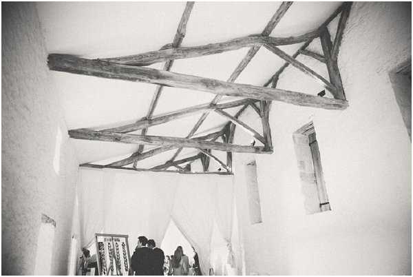A black-and-white wide-angle interior shot of a rustic barn or stone chapel venue, with exposed heavy wooden roof trusses forming a dramatic geometric pattern against white-washed walls and a vaulted ceiling. In the lower portion of the frame, a small group of people — including what appears to be a couple and several guests — stand near sheer white fabric draping that frames a doorway or altar area. A decorative framed display, possibly a seating chart or signage, is visible on the left side. The image has soft contrast with bright highlights on the walls and ceiling, and the composition emphasizes the architectural ceiling detail over the figures below. Potential venue feature image.