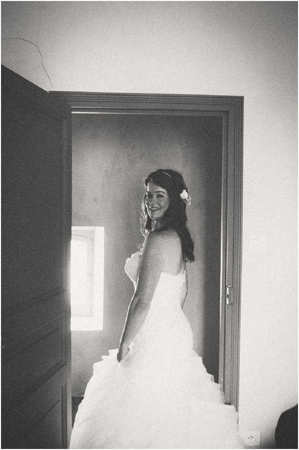A black-and-white portrait of a bride standing in a doorway, glancing back over her shoulder with a smile. She is wearing a strapless ball gown with a full, layered skirt, and has a small flower accessory in her dark hair, which falls loosely. The image is taken indoors, likely during the getting-ready phase, with natural light coming through a window in the background creating a bright, high-contrast effect. The composition is a medium full-length portrait shot with slightly grainy film-like tones.