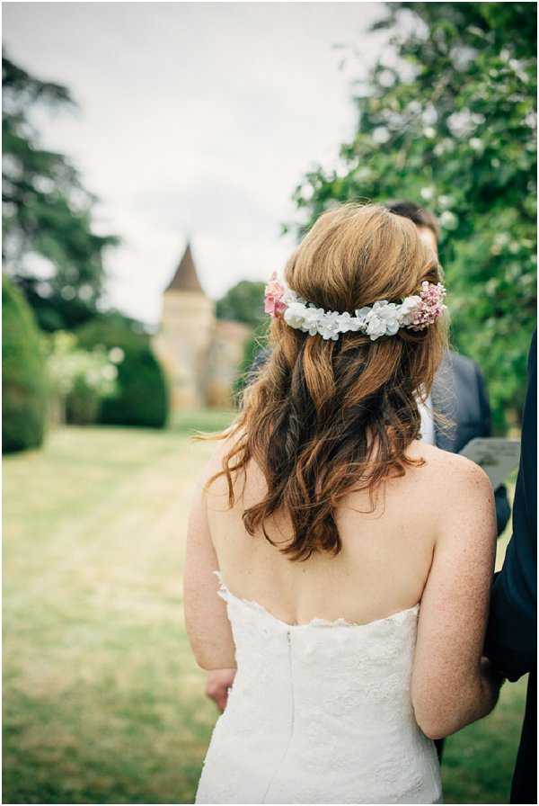 A portrait shot taken from behind of a bride standing outdoors on manicured grounds, with a groom partially visible to her right in a navy suit. The bride wears a strapless white lace dress and has auburn hair styled in a half-up, half-down arrangement with soft waves, adorned with a floral crown featuring white and dusty pink small blooms. In the background, a stone chateau tower with a pointed roof is visible through the trees, suggesting a French chateau setting. The overall styling has a romantic, garden-party feel with a bohemian touch from the flower crown.