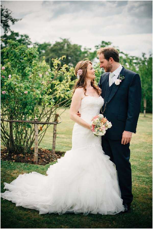 A couple portrait taken outdoors in a garden setting, with the bride and groom facing each other and smiling. The bride wears a strapless ivory fit-and-flare gown with a layered tulle skirt and a trailing train, and has a small blush pink floral hair accessory in her loose wavy hair; she holds a bouquet of soft peach, blush pink, and white mixed blooms. The groom wears a navy three-piece suit with a pale grey tie and a blush pink buttonhole flower. The shot is a medium full-length portrait with soft, slightly muted natural daylight and rose bushes visible in the background.