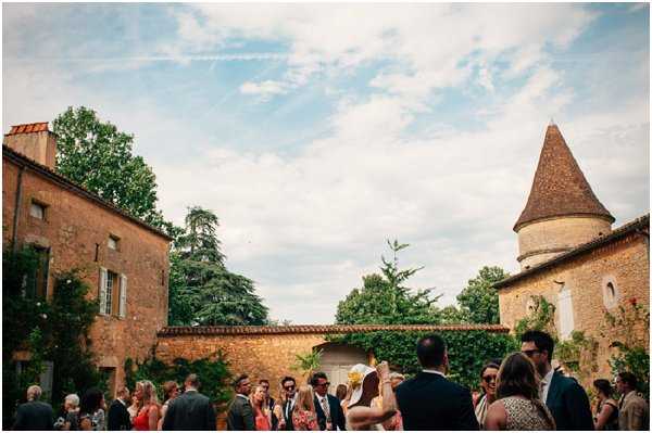 A cocktail hour or post-ceremony gathering taking place outdoors in the courtyard of a French chateau, with approximately 20–30 guests mingling in small groups. The venue features warm terracotta-roofed stone buildings on either side and a distinctive conical-roofed tower on the right, with ivy climbing the walls. Guests are dressed in summer attire including colorful dresses and suits, with one woman visible in a yellow hat. The shot is a wide-angle composition taken from ground level looking into the courtyard, capturing both the crowd and the full architectural surroundings. Potential venue feature image.