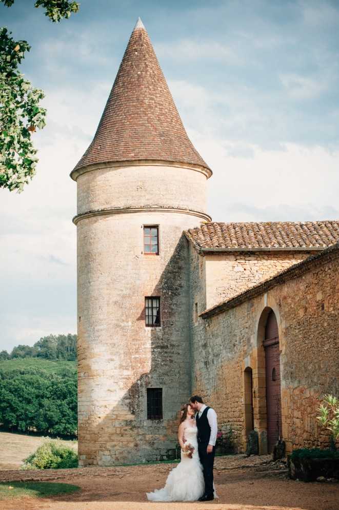 A couple portrait taken outdoors at the base of a round stone tower with a terracotta-tiled conical roof, part of a historic French chateau or manor complex with arched doorways and traditional clay tile roofing. The bride wears a white strapless ball gown with a full layered tulle skirt and holds a small bouquet, while the groom is dressed in a dark navy suit with a waistcoat and white shirt; the two are sharing a kiss. The shot is a wide portrait framing both figures small against the imposing tower architecture, with rolling countryside and tree lines visible in the background. Potential venue feature image.