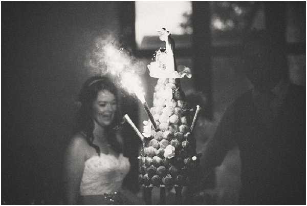 A black-and-white close-up shot of a traditional French croquembouche wedding cake in the foreground, decorated with small flowers and topped with a bride-and-groom cake topper, with sparklers lit and actively sparkling from the top. The bride, wearing a strapless dress and a hair accessory, stands smiling in the softly blurred background alongside another person, likely the groom. The image is taken at low light during an evening reception, with high contrast between the bright sparkler flash and the dark surroundings. The shallow depth of field keeps focus on the croquembouche while the couple remains out of focus behind it.
