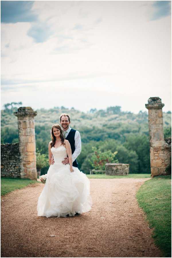 A couple portrait taken outdoors on a gravel path, with the groom standing behind the bride and his arms around her waist. The bride wears a strapless ivory ballgown with a layered, ruffled full skirt and holds a small bouquet with blush and ivory tones. The groom is dressed in a white shirt, navy vest, and dark trousers without a jacket. They are positioned between two tall, weathered sandstone pillar ruins, with a rolling wooded landscape visible in the background. The setting has a rustic, countryside feel typical of the Dordogne or Périgord region of France. The shot is a mid-length portrait with a slightly soft-focus background.