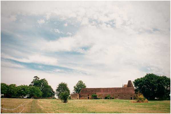 A wide-shot exterior view of a French rural wedding venue, featuring a long, low stone barn or farmhouse structure with terracotta-tiled roofing and a distinctive conical tower, set within open fields. The building spans the middle distance of the frame, surrounded by mature trees on either side. No people are visible in this image. The composition is a broad landscape shot with the venue occupying roughly the lower third of the frame. Potential venue feature image.