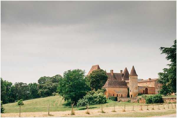 Wide exterior shot of a French chateau viewed from a distance, showcasing the full property set on a hillside. The building features classic medieval architecture with conical-roofed towers, terracotta brick walls, and multiple chimneys. No people or wedding activity are visible in this image; it serves purely as a venue establishing shot. Potential venue feature image.