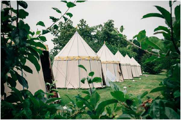 An outdoor wedding venue setup showing a row of large cream-colored canvas marquee tents with pointed peaks and yellow trim details, arranged across a lawn area. The tents appear to be traditional-style tipi or sperry-type structures used for wedding receptions or accommodation, with metal lanterns placed on the grass nearby. The shot is framed through foreground foliage, creating a wide environmental view of the tent arrangement. Potential venue feature image.
