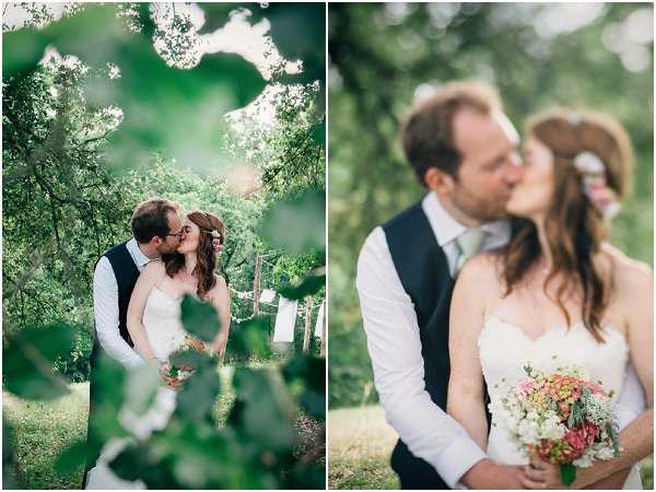 A two-image diptych of couple portraits taken outdoors in a wooded garden setting. On the left, a wide shot frames the couple kissing through leafy tree branches in the foreground, with bunting or hanging decorations visible in the background; the groom wears a navy waistcoat over a white shirt with a light grey tie, and the bride wears a strapless white fitted gown while holding a bouquet of coral, pink, and white mixed flowers including what appear to be hydrangeas and Queen Anne's lace. On the right, a close-up portrait shows the same couple kissing, with the bride's bouquet of coral and white blooms prominently visible and a small floral hair piece in her dark auburn hair. The styling is relaxed and rustic, with natural dappled light filtering through the trees in both shots.