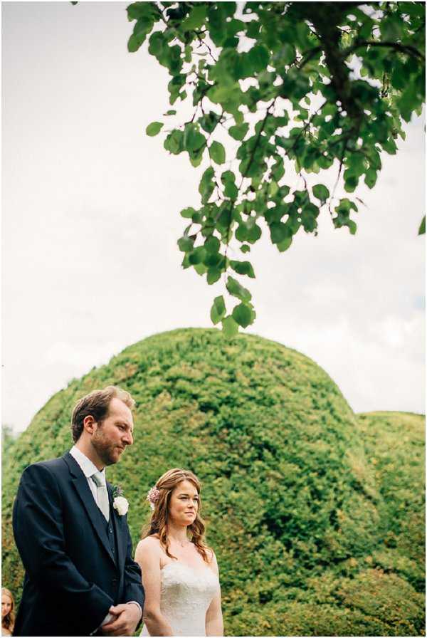 An outdoor wedding ceremony showing the couple standing together, captured in a medium portrait shot. The groom wears a dark navy suit with a grey tie and a white boutonnière, while the bride wears a strapless lace ivory gown and has a pale pink floral hair accessory. The setting features dramatically shaped rounded topiary hedges in the background, suggesting a formal garden, with a leafy tree branch framing the top of the composition. The overall styling is classic and understated.