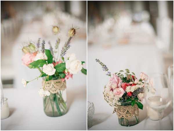 Close-up detail shot of two rustic wedding table centerpieces displayed side by side in a split-image composition. Each centerpiece consists of a glass mason jar wrapped with a lace or burlap trim, holding loose arrangements of cream and blush roses, coral blooms, lavender sprigs, thistle buds, and green foliage. Small glass votive candle holders are visible alongside the arrangements. The table is covered with a white linen, and the blurred background suggests a bright, airy indoor reception space styled in a rustic, vintage-inspired theme with a soft pastel color palette.