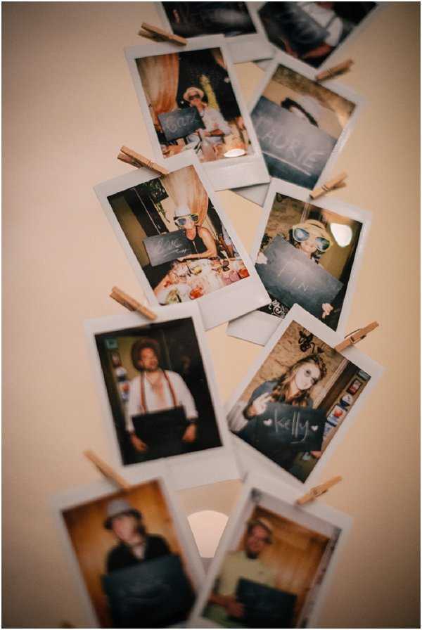 A close-up detail shot of a wedding guest book or photo display installation, featuring approximately 12–15 instant Polaroid-style photos clipped to strings or a surface using small natural wood clothespins. The photos show individual guests and small groups holding small chalkboard signs with their names written in chalk, including readable names such as 'Kelly' and 'Roxie.' Some guests are wearing novelty props such as oversized glasses and goggles. The display has a casual, boho-rustic aesthetic and appears to be mounted against a plain light-colored wall. The image is shot at a shallow depth of field, with the upper photos slightly blurred and the lower ones in sharper focus, giving a diagonal compositional flow.