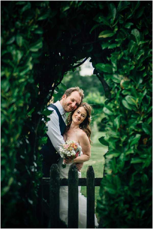 A couple portrait taken outdoors in a formal garden, with the bride and groom framed by a dense leafy hedgerow archway. The groom stands behind the bride, both smiling, with a dark wooden picket gate visible in the foreground. The bride wears a strapless ivory gown and holds a loose, wildflower-style bouquet featuring coral, pink, and white blooms. The groom wears a white dress shirt, dark navy waistcoat, and a teal tie. The composition is a close-to-medium portrait shot through the natural arch opening, with a garden path visible and softly blurred in the background, giving the image a framed, intimate quality.