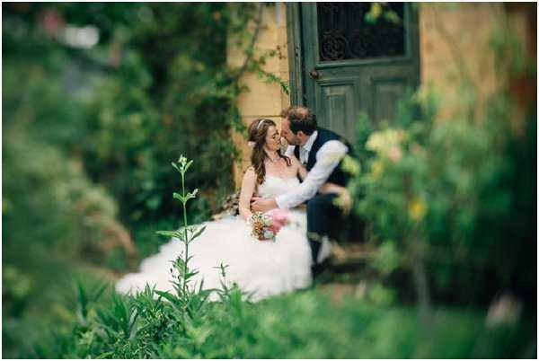A couple portrait taken outdoors in a garden setting, with the bride and groom seated in front of an aged green double door set into a yellow-painted wall. The bride wears a full-skirt white ballgown and holds a small mixed bouquet with warm-toned blooms. The groom is dressed in a dark navy vest, white dress shirt, and grey trousers, and leans in toward the bride. The shot is framed through soft foreground foliage, creating a shallow depth-of-field effect that partially blurs the couple while lush greenery fills the foreground and surrounds the doorway. The overall styling has a relaxed, garden-romantic feel.