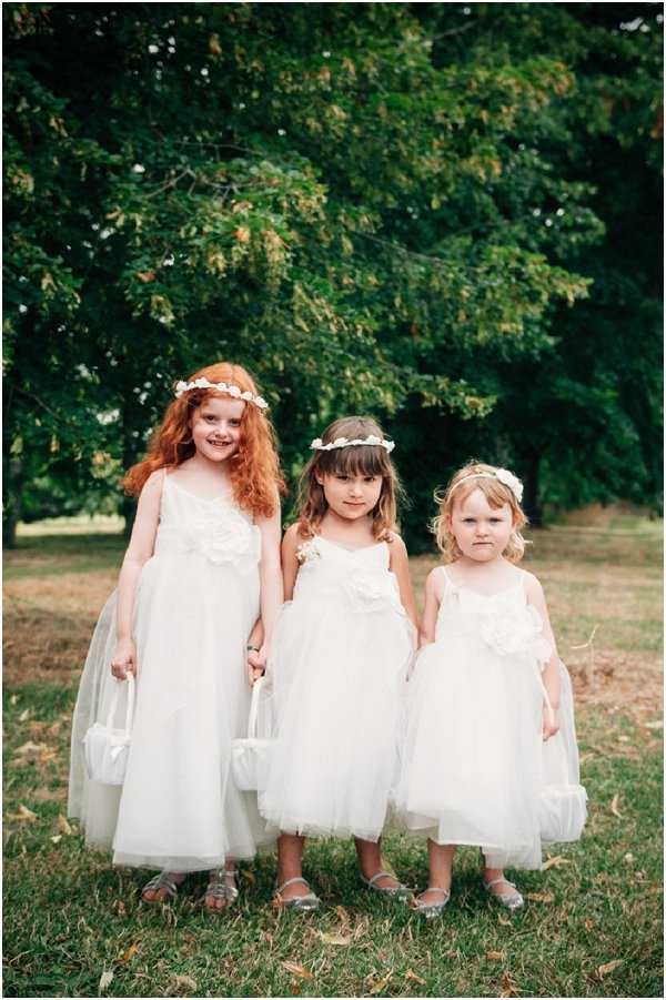 Three flower girls stand together outdoors on a lawn, posing for a portrait in front of large leafy trees. All three wear matching ivory tulle and satin spaghetti-strap dresses with floral appliqué detail at the bodice, and each carries a small ivory satin basket. Each girl wears a white floral crown headpiece. The children range in age from approximately toddler to around seven or eight years old. The styling is classic and cohesive with an all-ivory color palette. The shot is a mid-length portrait with soft, natural light.