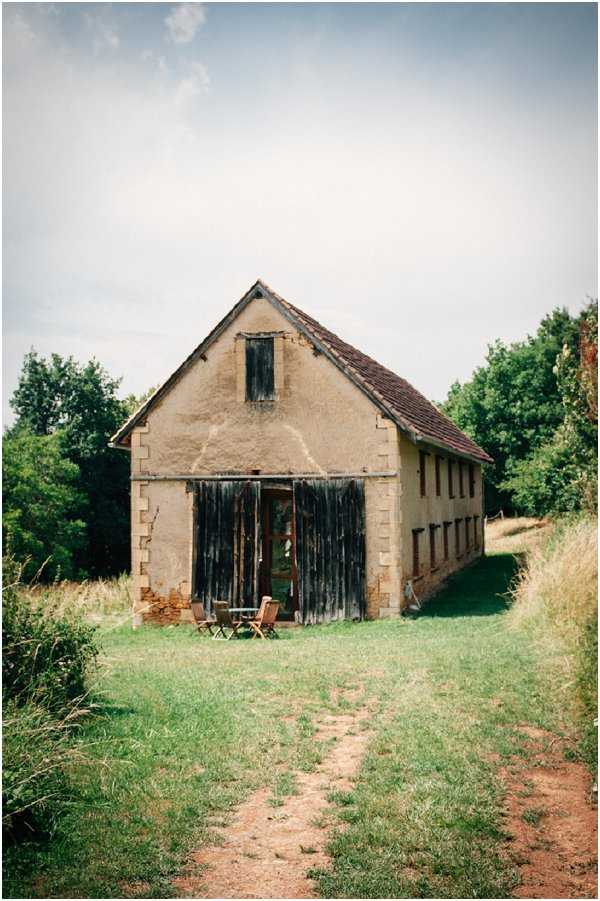 Wide shot of a rustic French barn or agricultural outbuilding with rendered stone walls, a terracotta tile roof, and weathered dark wooden doors standing open at the entrance. A small cluster of wooden folding chairs is visible just inside or in front of the open barn doors, suggesting the space is being considered or prepared as a venue. The building has a classic gabled facade with a small shuttered window near the peak and a row of smaller openings along the side wall. The setting is rural and outdoor, with a dirt path leading up to the entrance. Potential venue feature image.