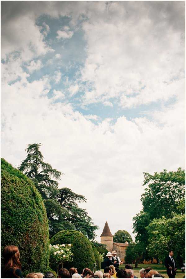 An outdoor wedding ceremony taking place in the grounds of a French chateau, visible in the background with its distinctive conical stone tower. The wide-angle shot is composed with the majority of the frame given to the open sky, placing the gathered guests, who number approximately 20-30 visible in the lower portion of the image, as a small element beneath manicured topiary hedges and large cedar trees. One guest wearing a straw hat and waistcoat stands out near the front. The overall aesthetic is relaxed and rustic, consistent with a countryside French chateau setting. Potential venue feature image.