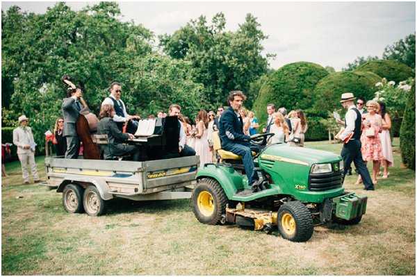 An outdoor wedding cocktail hour or reception activity taking place on a lawn garden, where a small live band of approximately four musicians — including a double bass player, a pianist at an upright keyboard, and at least one other musician — are performing on a flatbed trailer being towed by a green John Deere X740 ride-on lawn tractor driven by a man in a navy suit. Guests, numbering around 20–25 visible, are following or watching the mobile performance; attire is summer garden party style with women in colorful midi and mini dresses and men in suits and blazers. The garden setting features manicured topiary hedges in the background. The shot is a wide mid-distance photograph capturing the full scene in natural daylight with a slightly faded, film-style color treatment.