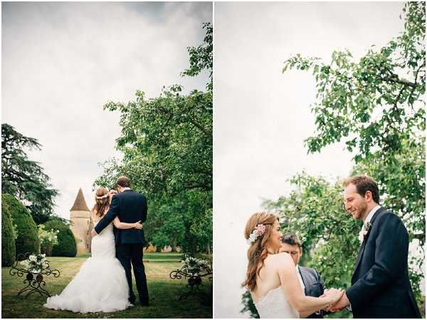 A diptych of two outdoor couple portraits taken in the grounds of a French chateau. In the left image, the bride and groom stand with their backs to the camera in a wide shot, embracing on a lawn with a stone chateau tower visible in the background and a wrought-iron decorative bench or arch with white florals nearby; the bride wears a fitted mermaid-style white gown with a ruffled skirt and has her hair half-up with a floral hair piece, while the groom wears a navy suit. The right image is a closer portrait showing the couple facing each other and smiling, with the bride's pink and white floral hair accessory clearly visible and the groom wearing a white boutonniere on his navy lapel. The overall styling is classic and romantic, with a natural garden setting, and the photography has a soft, slightly muted tone across both frames.