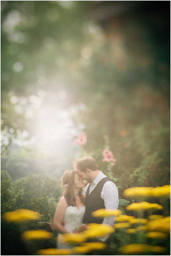 A couple portrait taken outdoors in a garden setting, with the bride and groom standing close together and touching foreheads in an intimate moment. The bride wears a strapless white gown and the groom wears a dark waistcoat over a white shirt with a light-colored tie. The foreground is filled with out-of-focus yellow marigolds, while soft pink flowers are visible in the background foliage. The image is shot with a very shallow depth of field and strong lens flare creating a soft, hazy glow, giving the portrait a painterly quality. The composition is a medium portrait with the couple centered in the frame amid lush garden greenery.