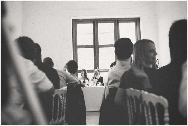 A black-and-white wide shot taken during an indoor wedding reception, showing a couple seated at a head table in the background, framed by a large multi-pane window that provides strong backlight. The high contrast image renders the foreground guests as dark silhouettes, while the bride and groom are faintly visible at the table in the mid-ground. Rows of what appear to be white Chiavari chairs with dark sashes are visible on either side of the aisle-like space. The venue interior features plain white walls, suggesting a converted barn or chateau outbuilding with a rustic aesthetic.