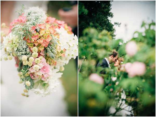 A two-image composition featuring a close-up detail shot on the left and a couple portrait on the right. The left image is a close-up of a bridal bouquet held against a white dress, composed of coral and blush hydrangeas, cream white hydrangeas, yellow-green billy balls, white Queen Anne's lace, pink larkspur, and trailing fern and greenery in a loose, garden-style arrangement. The right image shows the bride and groom sharing a close moment, nearly kissing, photographed through a foreground of blush pink hydrangea blooms and dense green foliage, creating a soft, framed portrait effect. The groom wears a navy suit and the couple are surrounded by lush garden plantings, suggesting an outdoor garden or chateau grounds setting.