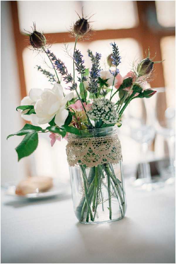 Close-up detail shot of a rustic wedding table centerpiece featuring a clear glass mason jar wrapped with a cream lace trim and filled with water. The arrangement includes a white garden rose, blush pink rosebuds, purple lavender stems, white scabiosa, and dried nigella seed pods. The jar sits on a white tablecloth, with blurred glassware and a small plate visible in the background, suggesting an indoor reception setting. The overall styling is rustic-boho with a wildflower, foraged aesthetic.