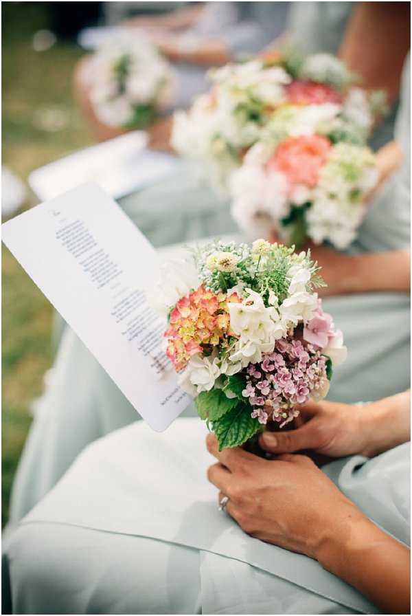 Close-up detail shot of bridesmaids seated during an outdoor wedding ceremony, each holding small round bouquets and printed ceremony programs. The bridesmaids are wearing pale sage/mint chiffon dresses. The foreground bouquet contains white hydrangeas, mauve hydrangeas, coral and yellow toned flowers, and fresh mint leaves, while the bouquet in the background includes coral roses, white hydrangeas, and greenery. The styling is soft and garden-inspired with a mixed pastel palette of coral, white, and mauve florals complementing the sage dresses. The shallow depth of field draws focus to the nearest bouquet and program.