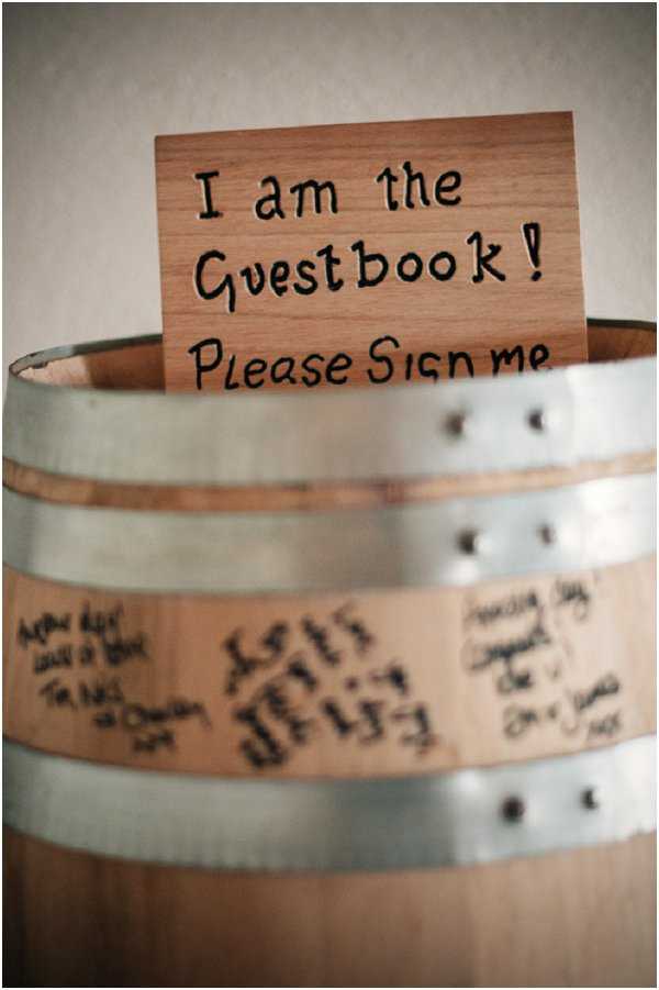Close-up detail shot of a rustic wedding guestbook concept using a wooden wine barrel with silver metal hoops. A small rectangular wooden sign with hand-burned lettering reading 'I am the Guestbook! Please Sign me' is propped inside the open top of the barrel. Guests have signed their names and written messages directly onto the natural wood surface of the barrel in black marker, with several signatures and handwritten notes visible around the lower half. The styling is rustic and vineyard-inspired, consistent with a French wine country wedding aesthetic.