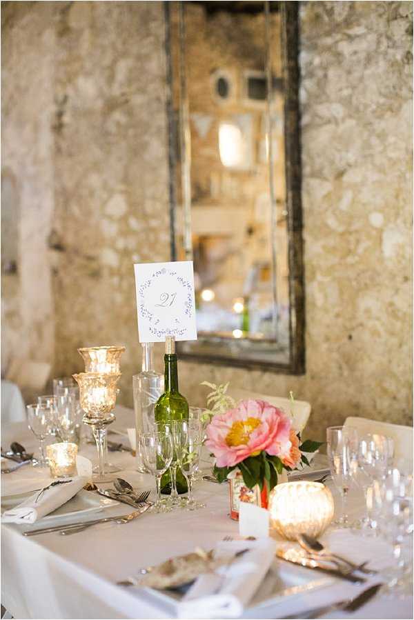 Close-up detail shot of a wedding reception table setting inside a stone-walled venue. The table is dressed with a white linen cloth and set with silver cutlery, crystal glassware including champagne flutes and wine glasses, and white cloth napkins. A green wine bottle holds a printed table number card (number 21) with a delicate wreath illustration. Centerpiece florals include a large coral-pink peony with foliage in a low arrangement. Candlelight comes from mercury glass votive holders and a taller mercury glass pedestal candle holder, giving the table a warm glow. An antique-framed mirror with a distressed dark frame hangs on the exposed stone wall in the background. The overall decor palette is white, gold, and coral-pink with rustic-chic styling.