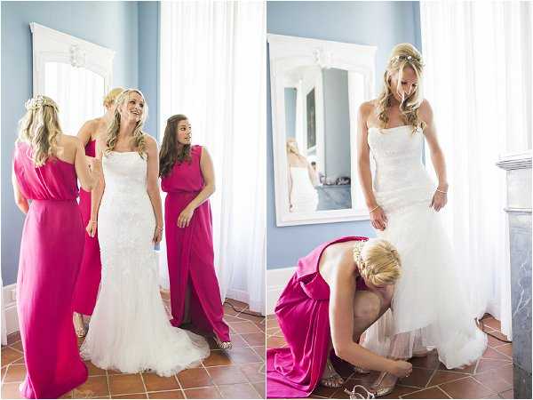 A getting-ready scene shown as a two-image side-by-side composition, set in an indoor room with blue-painted walls, terracotta tile flooring, and a large ornate white-framed mirror. On the left, the bride stands in a strapless lace fit-and-flare white gown while two bridesmaids in fuchsia pink floor-length dresses assist her and share a laugh; floral hair accessories are visible on at least one bridesmaid. On the right, a close-up portrait shows the same bride in her white lace gown looking down as a bridesmaid in a fuchsia dress kneels at her feet to fasten her strappy heeled sandals, with the mirror reflecting the scene behind them. Both images have a bright, airy feel with natural light, and the overall styling theme is classic with a bold fuchsia bridal party color palette.