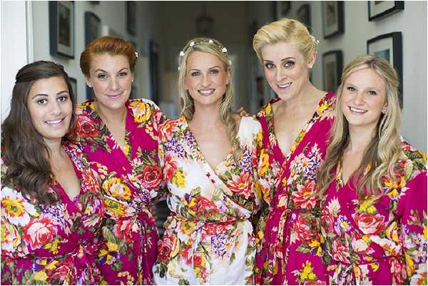 A getting-ready scene showing a bride and four bridesmaids posing together indoors, likely in a hallway or corridor with framed pictures visible on the wall behind them. The bride wears a white satin robe and a delicate floral crown of small white flowers in her hair, while the four bridesmaids wear matching magenta floral print robes featuring large pink, yellow, and purple rose patterns on a white background. All five women have their hair and makeup done, with the bride wearing a side braid. The group is smiling directly at the camera in a close portrait-style shot.