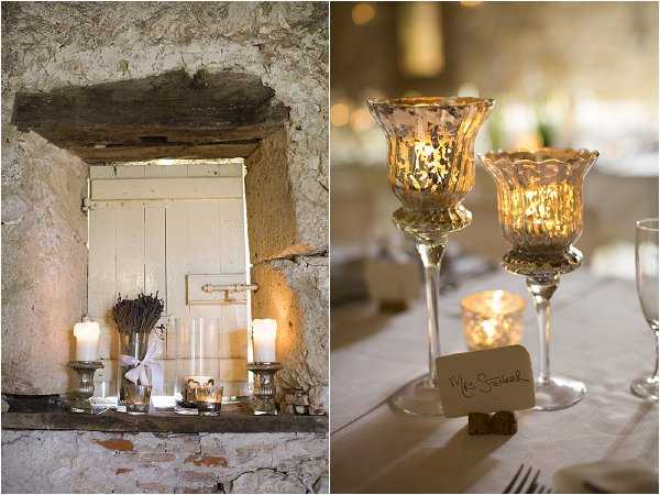 A split image showing two reception decor detail shots from what appears to be a rustic French venue. The left image shows a stone wall alcove with a white wooden shutter, styled with white pillar candles in glass hurricane holders and a small bundle of dried lavender tied with a white ribbon, creating a warm candlelit vignette. The right image is a close-up of a reception table setting featuring two tall mercury glass pedestal candle holders in gold tones, a place card reading 'Mrs. Steineck' propped against a wine cork, and glassware visible in the background, all bathed in warm candlelight. The overall decor palette is warm gold and ivory with rustic French countryside styling.