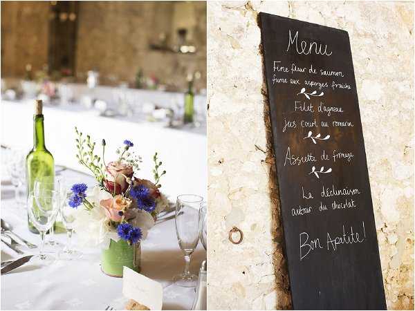 A detail-focused split image showing two close-up shots from a wedding reception set in a rustic stone-walled venue. On the left, a reception table is dressed with a white linen cloth and features a small floral centerpiece of bright blue cornflowers, blush and coral roses, and delicate white blooms in a mint green vessel, alongside green wine bottles used as casual decor and standard glassware. On the right, a hand-lettered chalkboard menu sign leaning against a limestone stone wall reads 'Menu' with four courses written in French chalk script, ending with 'Bon Appétit!' — the courses include fine smoked salmon with fresh asparagus, lamb fillet with rosemary jus, a cheese plate, and a chocolate dessert. The overall styling is relaxed and rustic with a wildflower-inspired color palette of blue, blush, and coral.