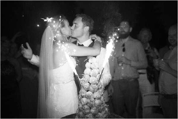 A black-and-white image of the bride and groom sharing a kiss behind a traditional French croquembouche wedding cake topped with sparklers, which are actively burning and creating bright light streaks in the frame. The bride wears a lace gown and a long veil, while the groom is in a suit with a waistcoat. Several guests are visible in the background watching the moment, with the scene set outdoors at night. The image has a grainy, high-contrast quality typical of nighttime film or high-ISO photography, giving it a slightly soft focus. Mid-range portrait shot.