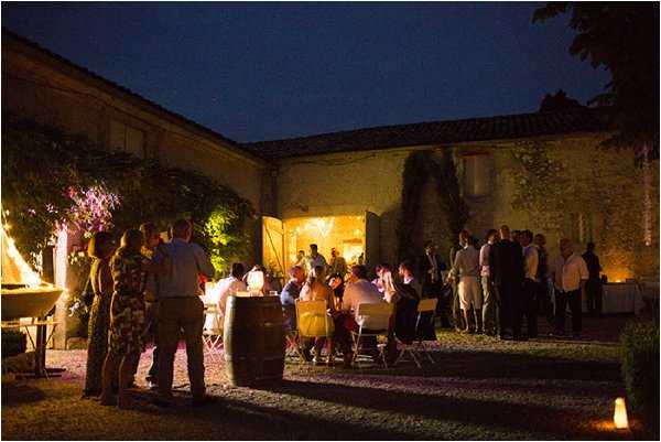 An outdoor evening reception taking place in the courtyard of a French stone farmhouse or rural estate, with approximately 30–40 guests gathered in groups across the gravel space. Some guests are seated at round tables with white linens while others stand in conversation clusters near the building's entrance, which is warmly lit from within. Wooden wine barrels are used as decorative and functional elements, and small ground-level candle lanterns line the pathway in the foreground. The warm amber uplighting on the stone building façade contrasts with the deep blue of the night sky, giving the scene a warm, rustic atmosphere consistent with a Provençal or Southwest French countryside aesthetic. Wide-angle shot capturing the full courtyard setting. Potential venue feature image.